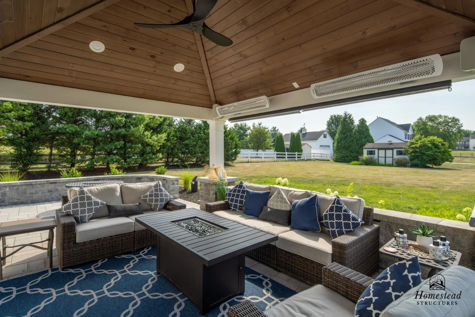 Outdoor patio with beige sectional sofa with blue and beige patterned pillows, a black fire pit table, a small side table with decorative jars, under a wooden ceiling with ceiling fans, surrounded by green lawn, shrubs, trees, and a white fence.