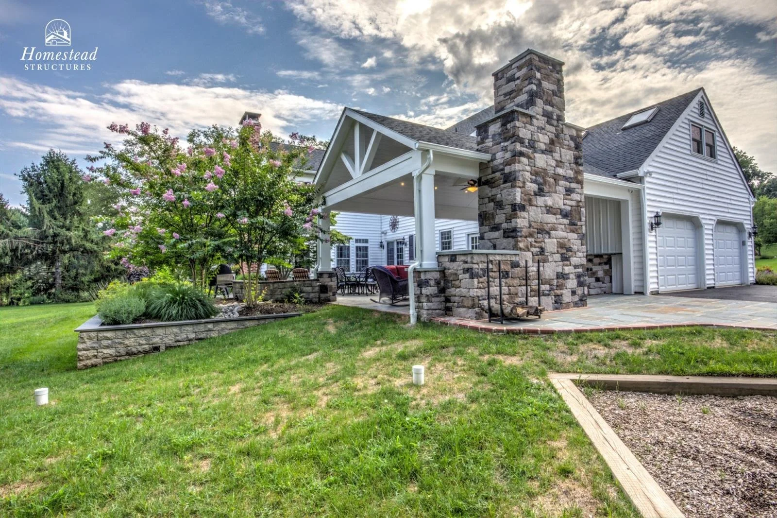 A house with white siding, a stone chimney, and a two-car garage. There is a covered patio with outdoor furniture, a flowering tree, and a well-maintained lawn.