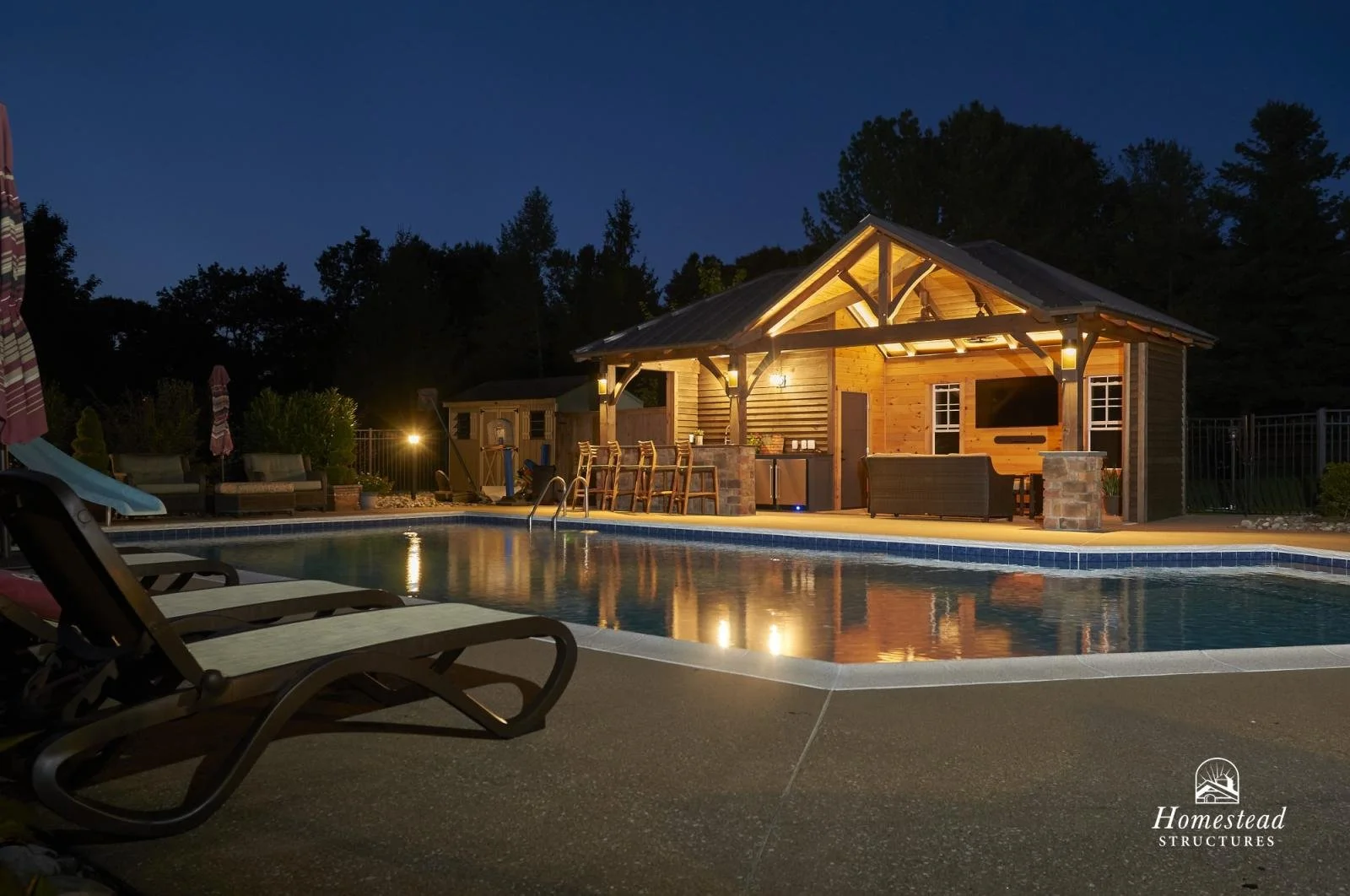 Nighttime view of a backyard pool area with a wooden cabana featuring a roof and lighting, outdoor furniture including lounge chairs, and a small shed in the background, surrounded by trees.