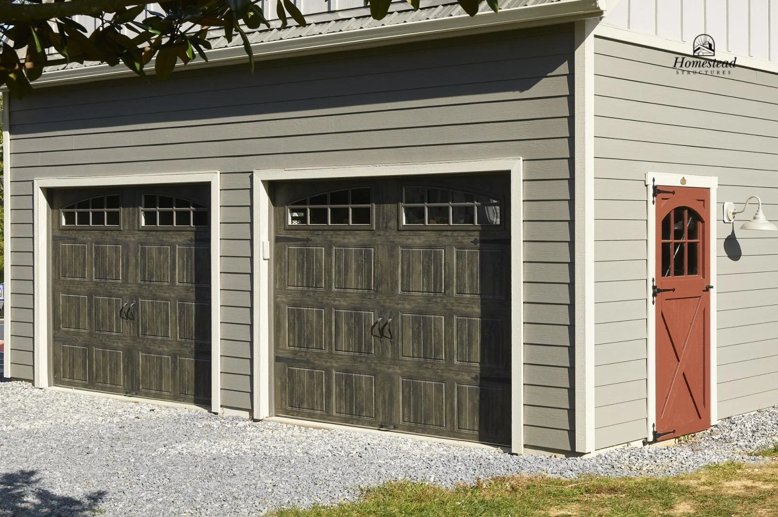 A two-car garage with dark brown wood panel doors and a small side door with a red barn door design on a light gray house. There is a white exterior light fixture on the right side.