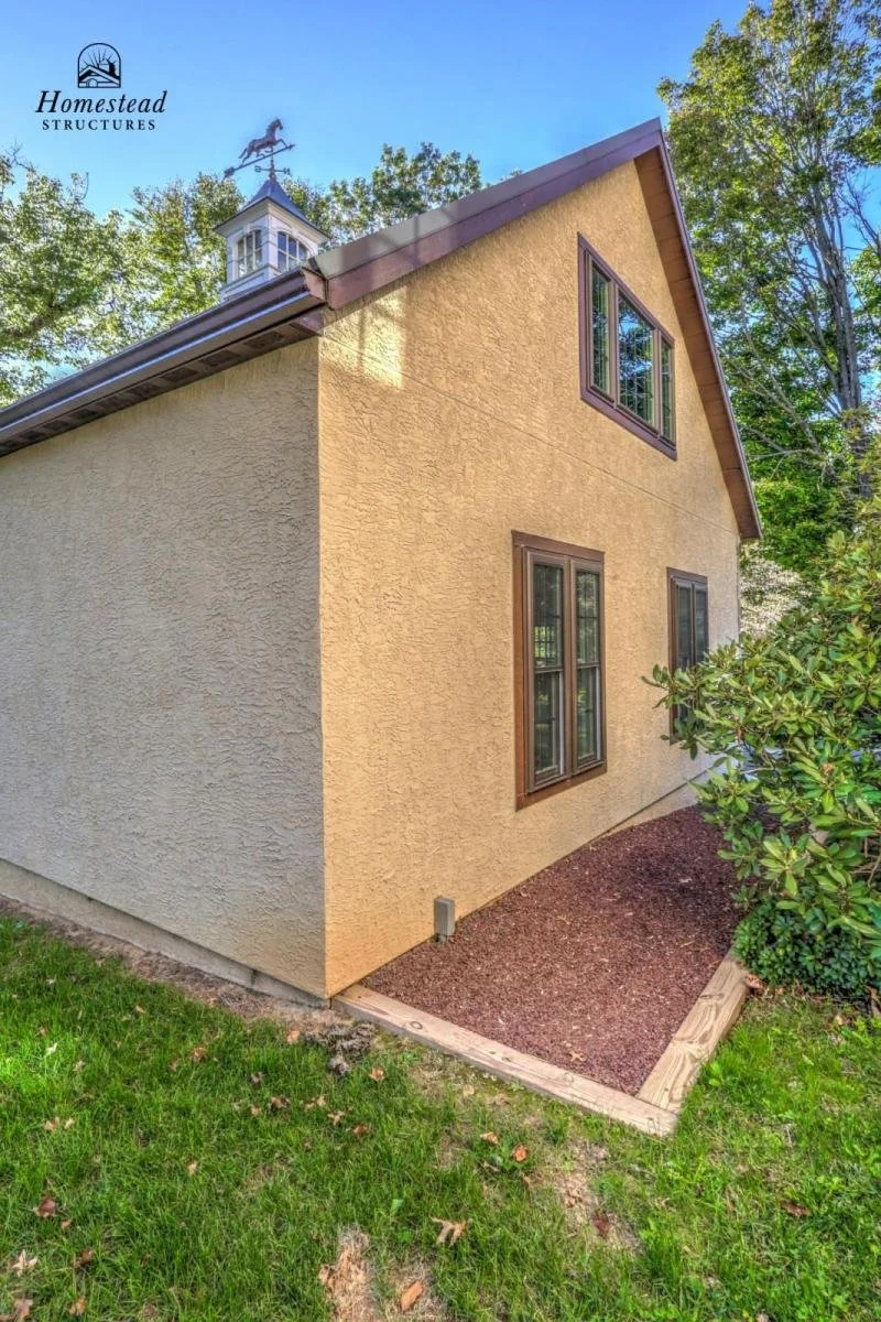 Side view of a newly built house with a sloped roof, several windows with brown frames, yellow stucco exterior walls, and a small landscaped area with mulch and a wooden border. The house is surrounded by green trees and grass.
