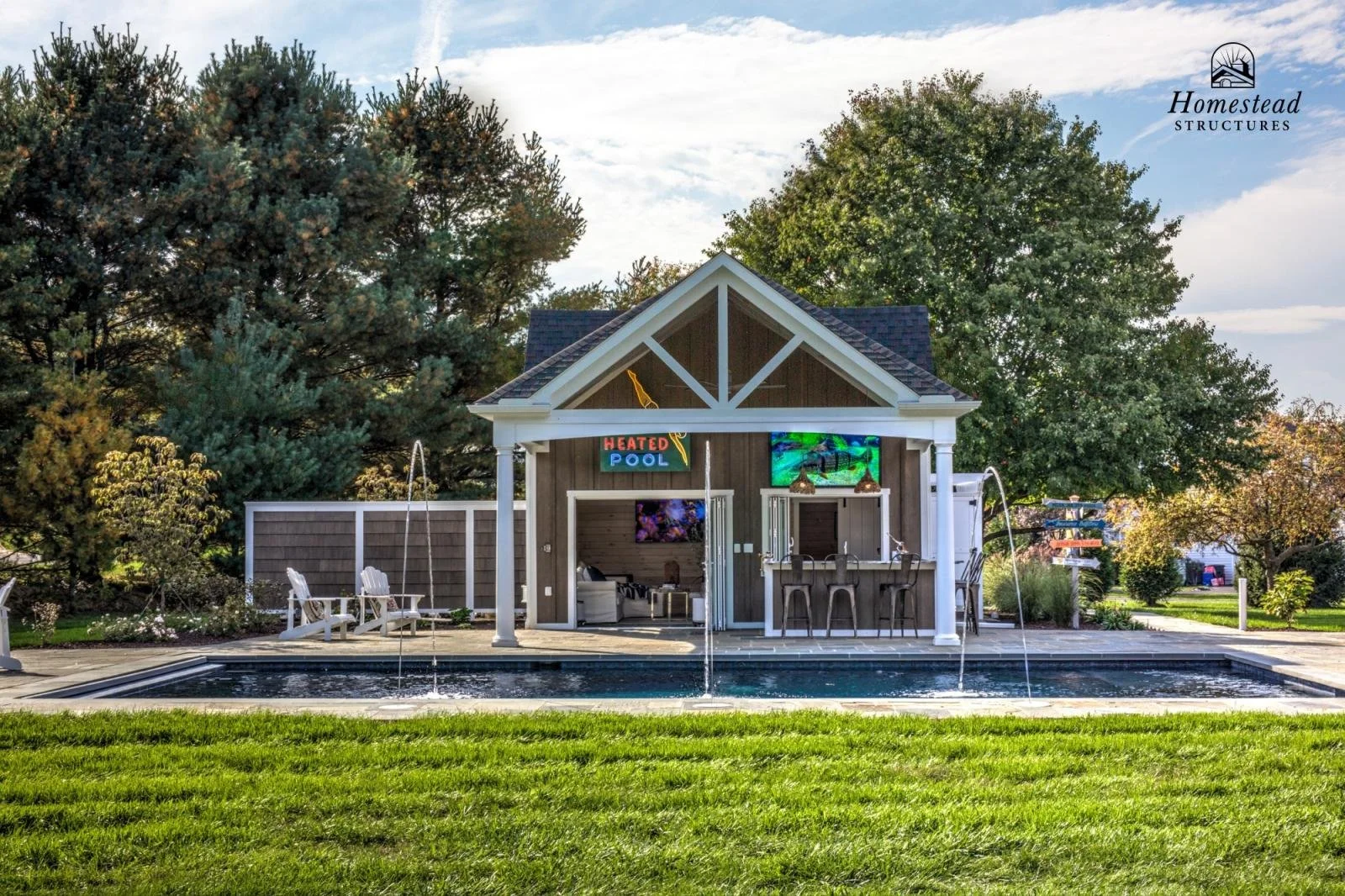 Outdoor pool area with a small pool, surrounded by green grass, trees, and a wooden cabana with a bar inside. The cabana has a neon sign that says 'Heated Pool' and two televisions. There are chairs and outdoor furniture.