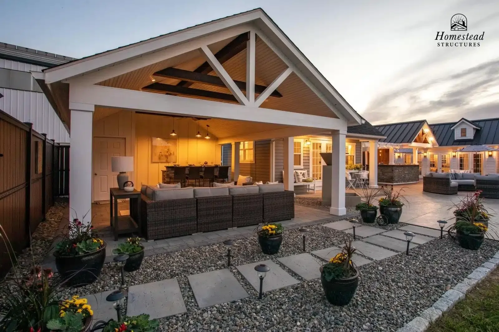 Exterior view of a modern, well-lit home patio at dusk with outdoor seating, potted plants, and decorative lighting.