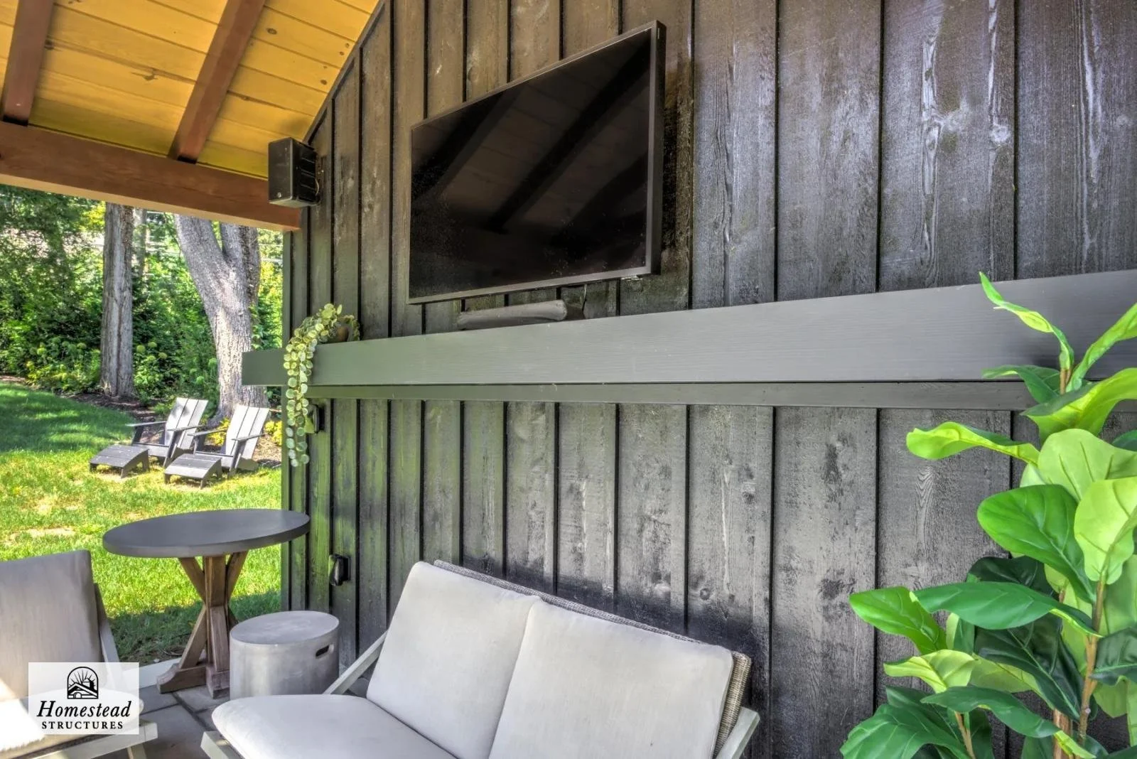 Outdoor patio with cushioned chairs, a side table, a large flat-screen TV mounted on a dark wooden wall, and lush green trees and grass in the background.
