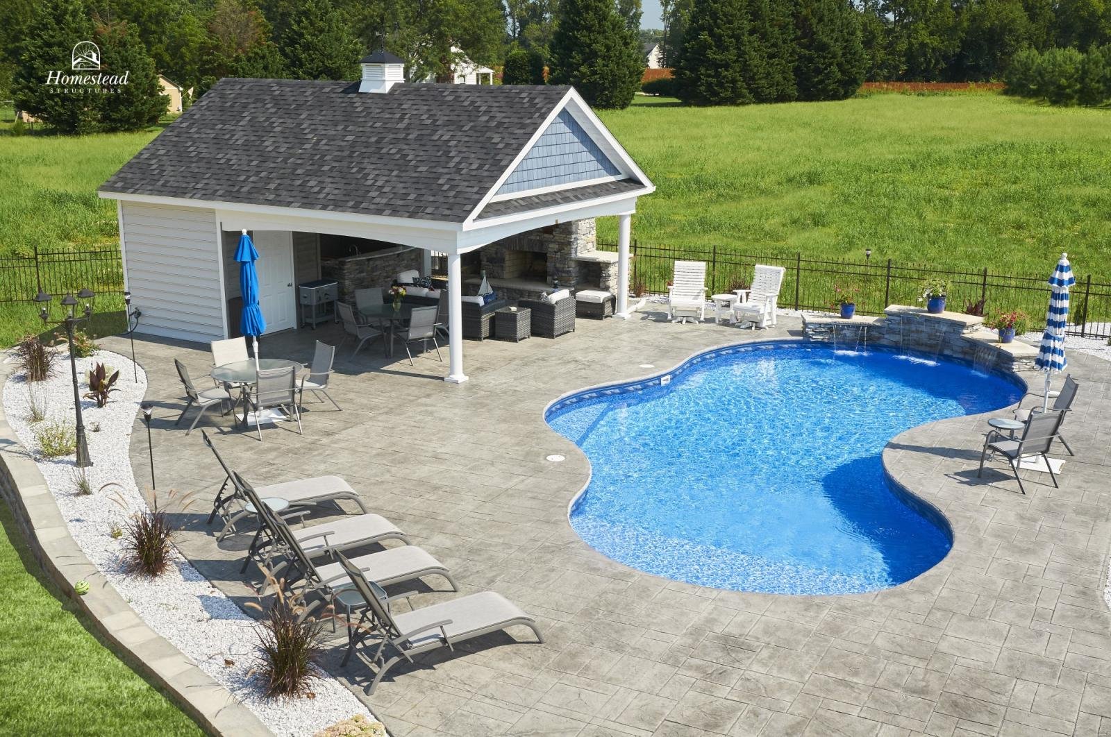 A backyard patio with a kidney-shaped blue pool, surrounded by lounge chairs, a seating area with an umbrella, and a poolside building with a roof, outdoor furniture, and a stone fireplace. Green lawn and trees are in the background.