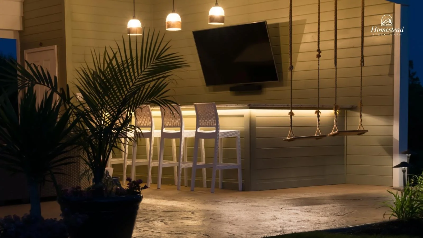 Outdoor patio with a green wall, hanging pendant lights, a mounted flat-screen TV, a bar with three barstools, and a wooden swing set hanging from the ceiling. Potted plants and patio flooring are also visible.