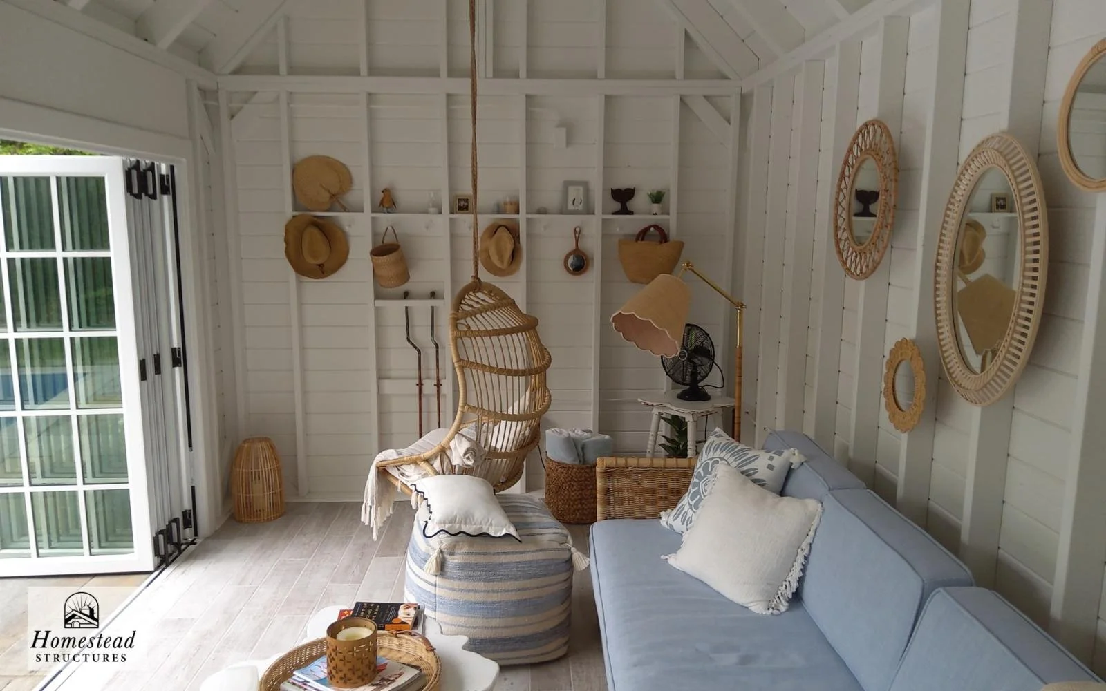 A cozy sunroom with white wooden walls and ceiling, decorated with wicker hats, baskets, and mirrors, featuring a light blue sofa with white pillows, a striped pouf, a small white side table with a vintage fan and lamp, and a door leading outside.