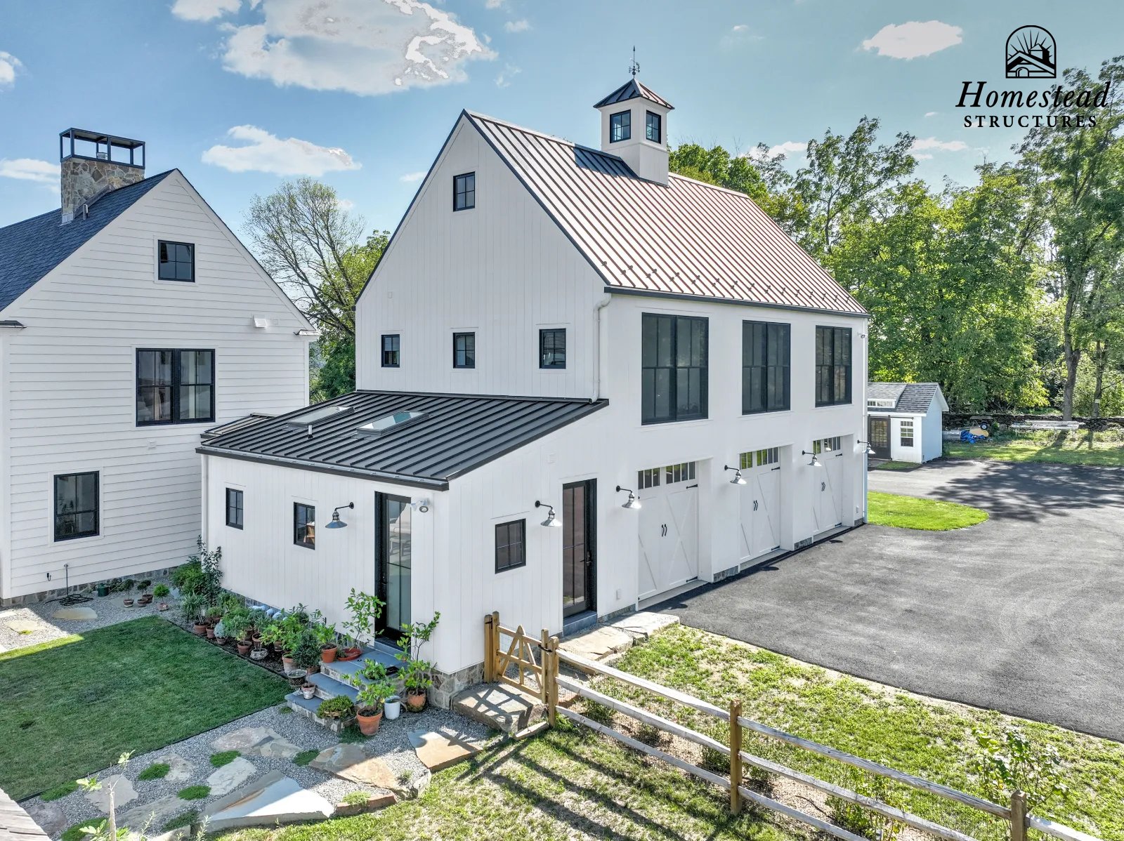 A modern white barn with black metal roof and large windows, surrounded by trees and a gravel driveway with a small fenced yard and garden.