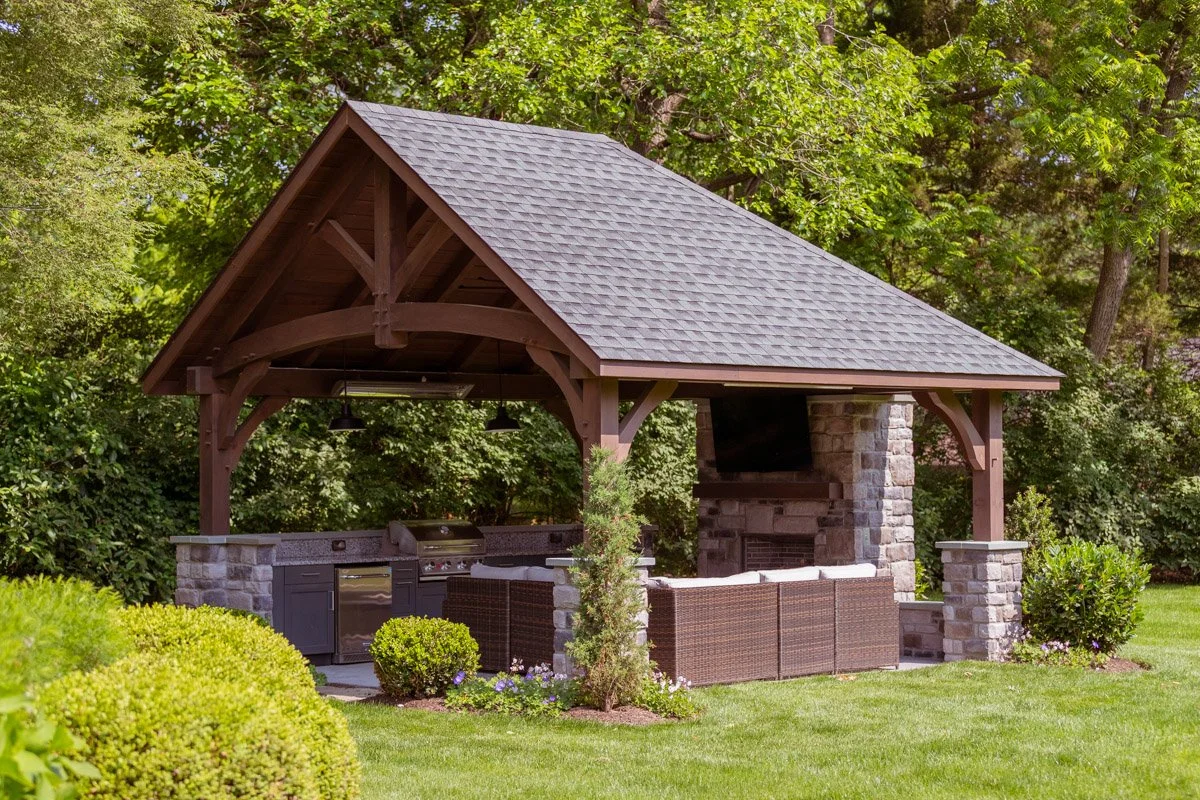 Outdoor backyard kitchen with a stone fireplace, a TV, and a shaded wooden roof, surrounded by greenery.