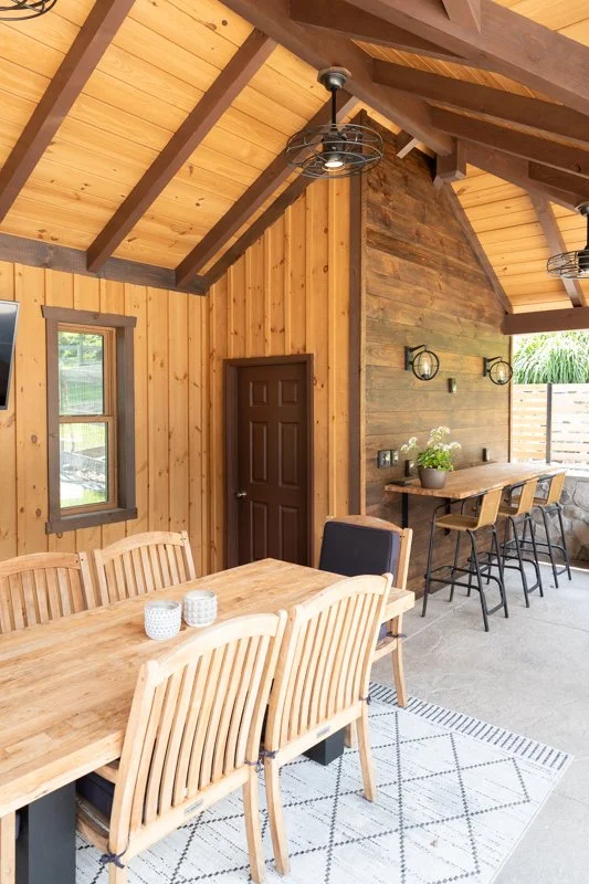 Interior of a wooden outdoor dining and bar area with a large dining table, eight chairs, and a bar counter with four stools, decorated with potted plants and wall-mounted lights.