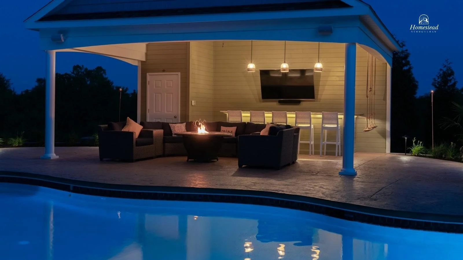 Outdoor patio area with a swimming pool, a fire pit surrounded by outdoor seating, a TV on the wall, bar stools, and hanging swing, illuminated at dusk.