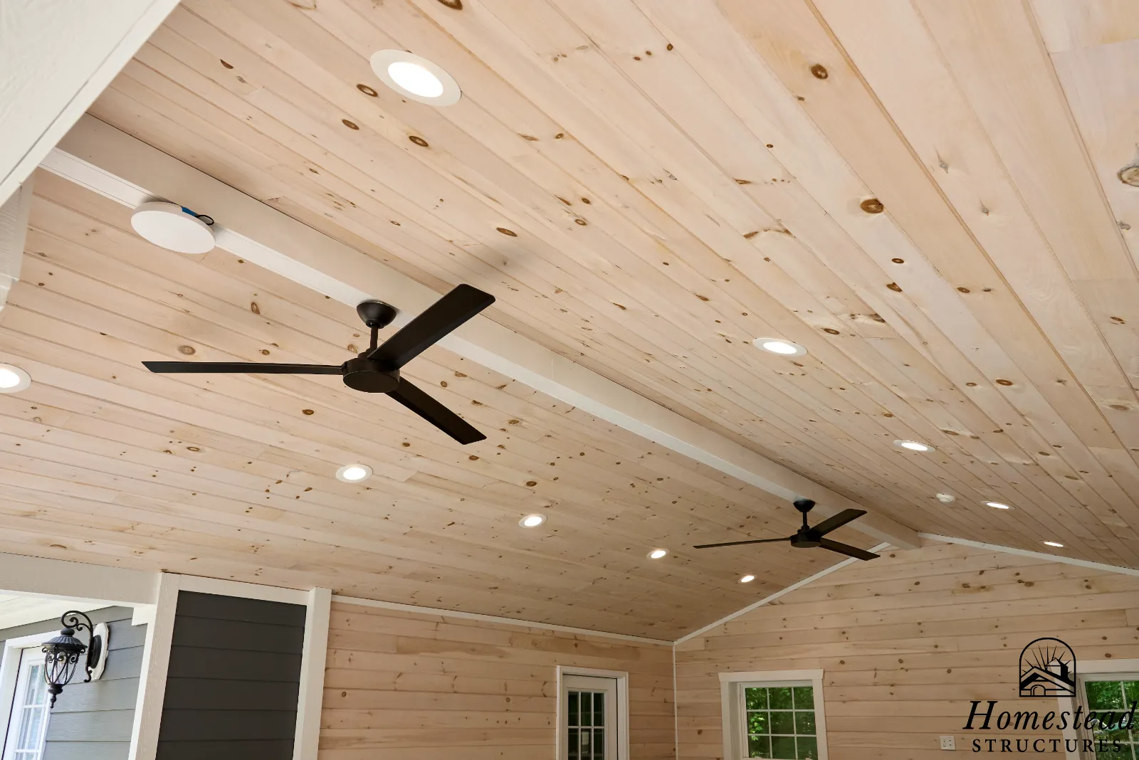Interior of a room with wood-paneled ceiling, black ceiling fans, recessed lighting, and walls with white trim and windows.