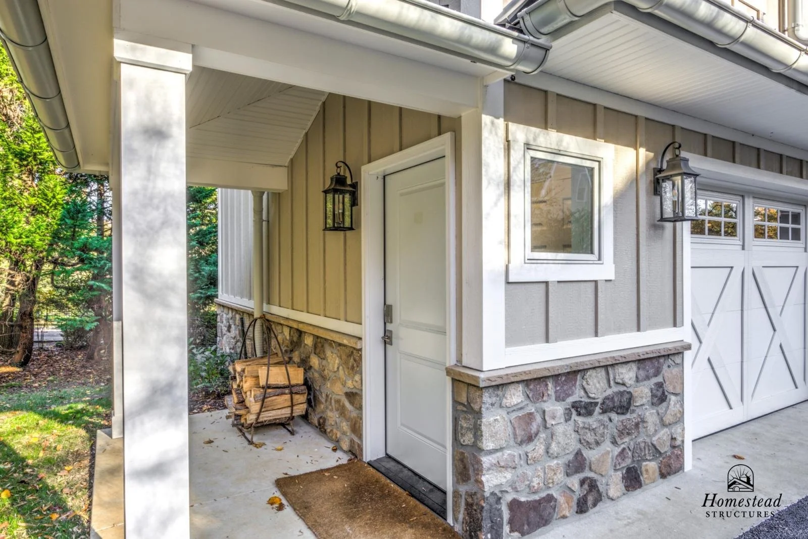 Front porch area of a house with stone and siding exterior, a white door, and two black lantern-style light fixtures, with a stack of firewood on a metal rack next to the house.
