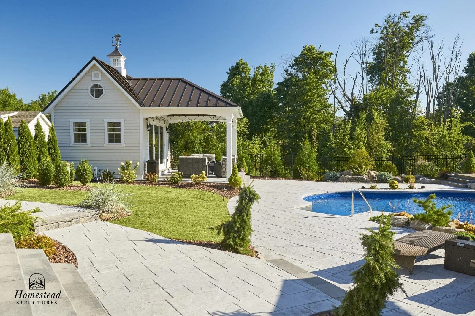 A backyard with a small white house featuring a covered porch, surrounded by manicured lawn and shrubs. There is a swimming pool with a metal ladder, and outdoor furniture, including lounge chairs, on the paved patio area, with trees and greenery in 