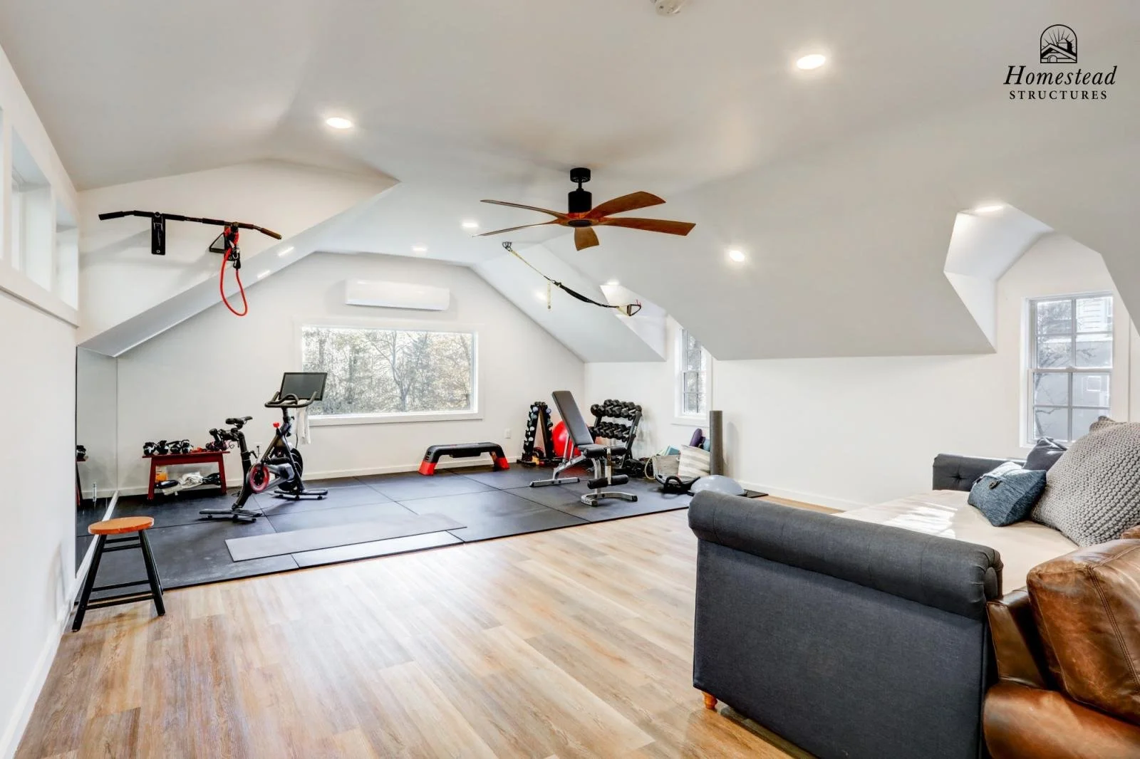 Home gym in an attic with exercise equipment, weights, a bench, a stationary bike, exercise mats, and a window with trees outside.