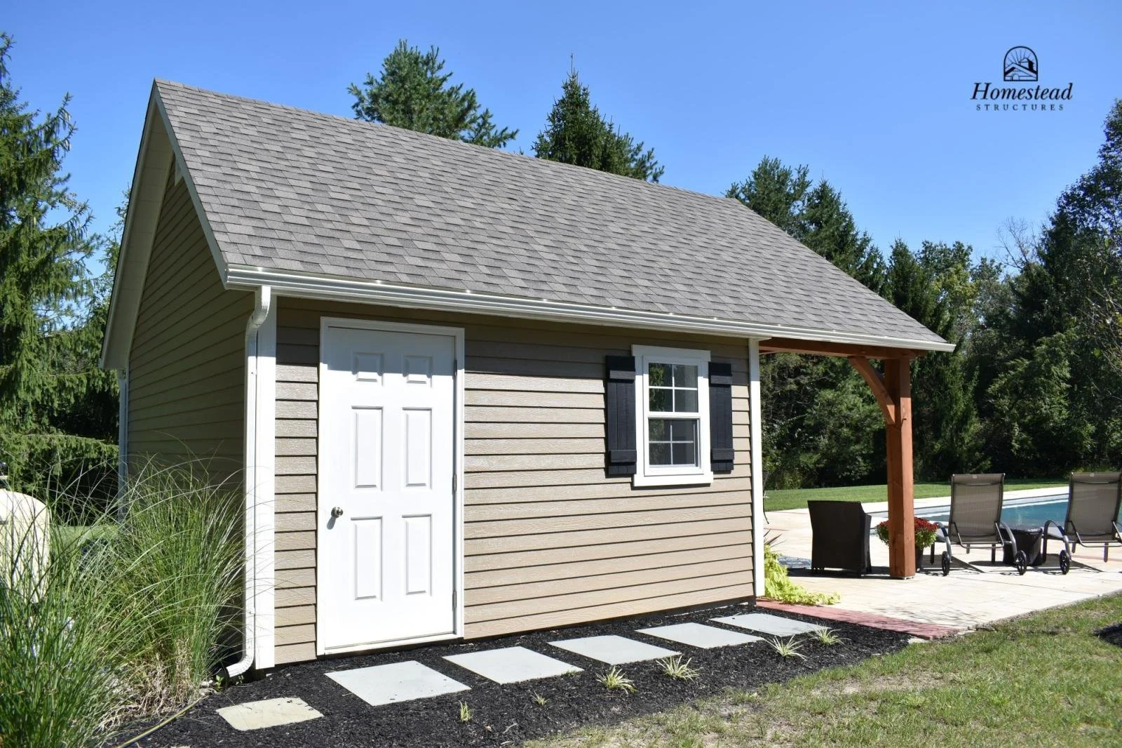 A small beige shed with a white door and a window with black shutters, located next to a swimming pool and patio chairs, with trees in the background under a clear blue sky