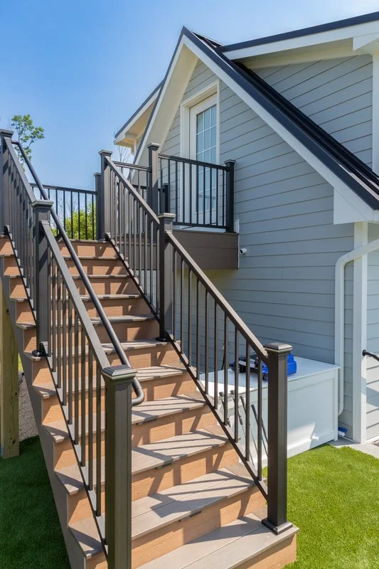 Outdoor wooden staircase with black metal railing leading to the second floor of a light gray house with white trim and a black roof.