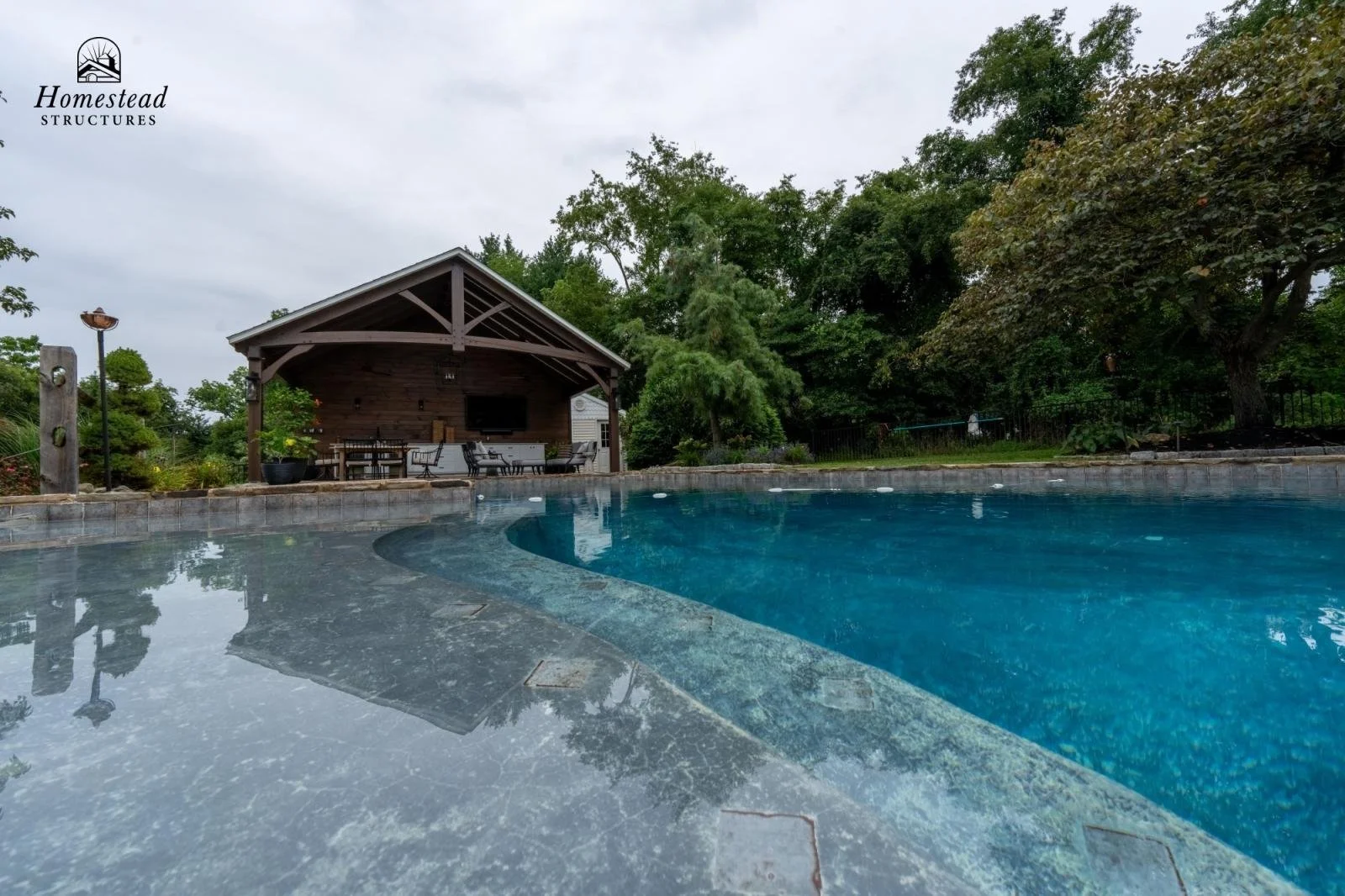 Residential backyard with a swimming pool, covered outdoor patio with a TV, dining table, and chairs, surrounded by trees and greenery.