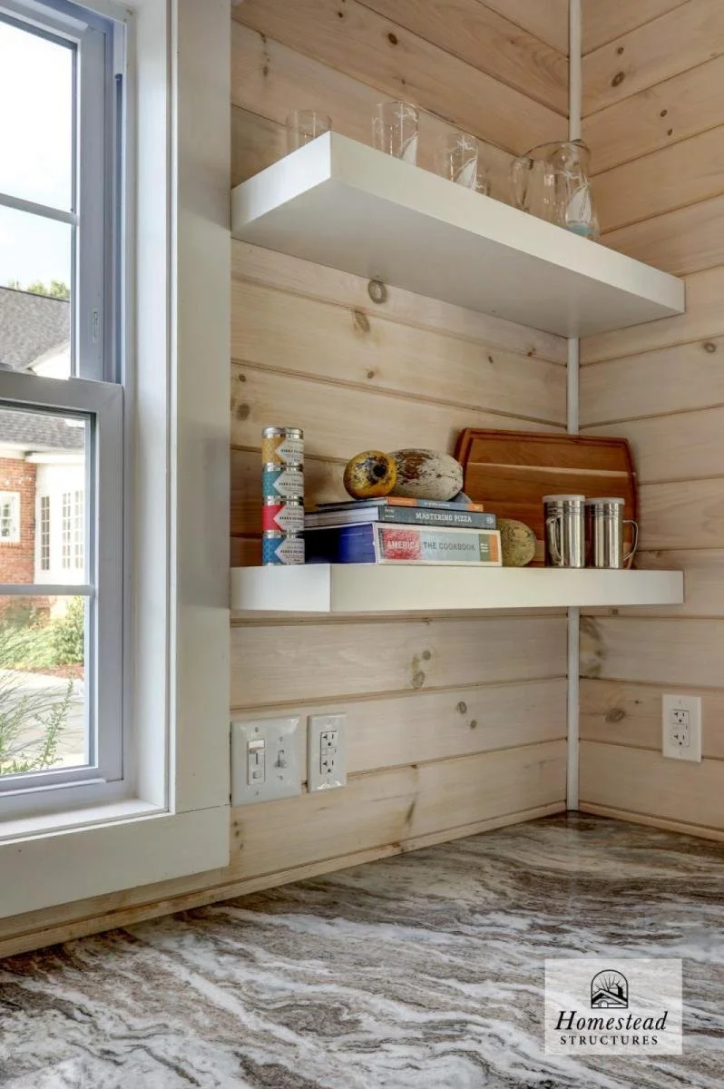 Interior view of a kitchen corner with light wooden wall panels, a window to the left, granite countertop, and two white floating shelves holding glass jars, books, decorative gourds, and metal containers. There are electrical outlets and switches on
