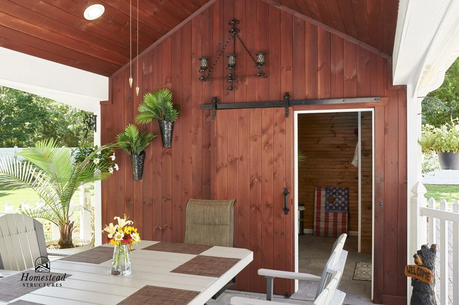 Patio area with a farmhouse-style wooden wall and ceiling, decorated with hanging plants in black pots, a black metal chandelier, and a sliding barn door leading to a wooden interior room with an American flag-themed chair. There is an outdoor dining