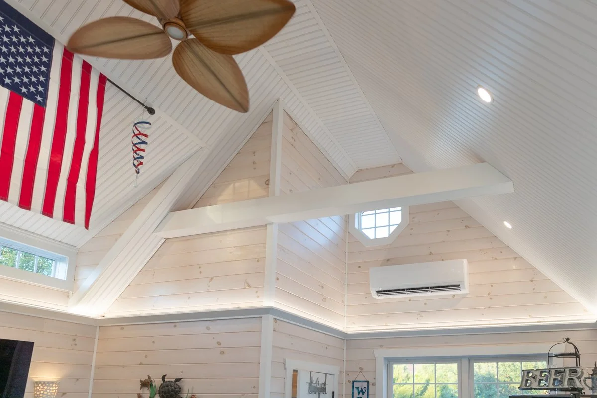 Ceiling of a bright, white-paneled room with a wooden fan, American flag, and triangular window.