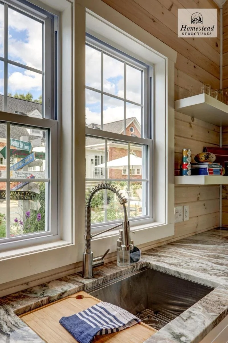 Inside a house kitchen with a granite countertop, a stainless steel sink, and a modern spring-coiled faucet. Two large windows above the sink reveal a suburban neighborhood with houses, trees, and a partly cloudy sky. A striped dish towel rests on a 
