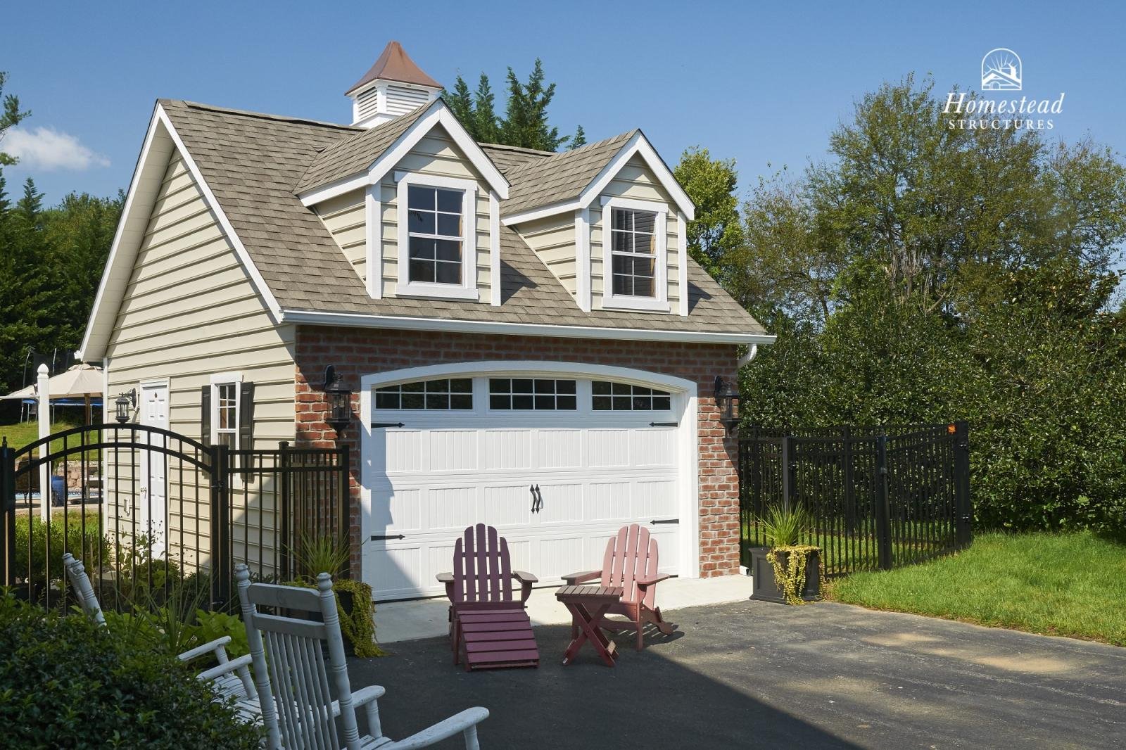 A small garage with a white door, brick and siding exterior, and two small dormer windows on a gabled roof. In front, two Adirondack chairs in pink and one in purple are positioned on a paved driveway. A black fence surrounds the area and there are t
