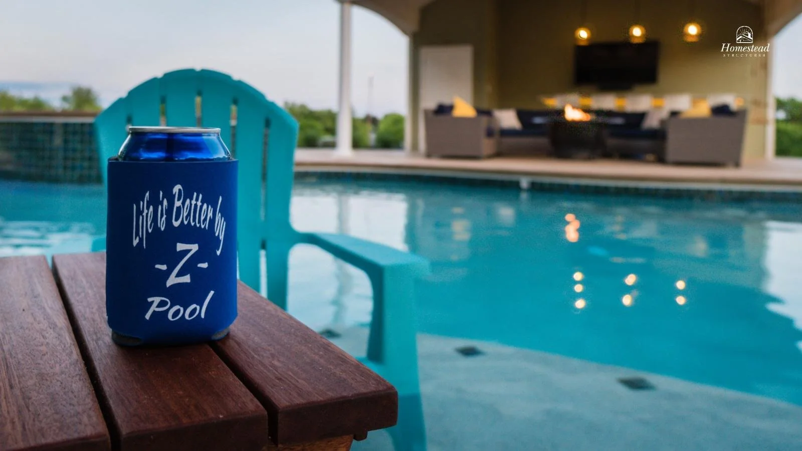 Blue can with a koozie reading 'Life is Better by -Z- Pool' on a wooden table next to a pool with outdoor furniture in the background.