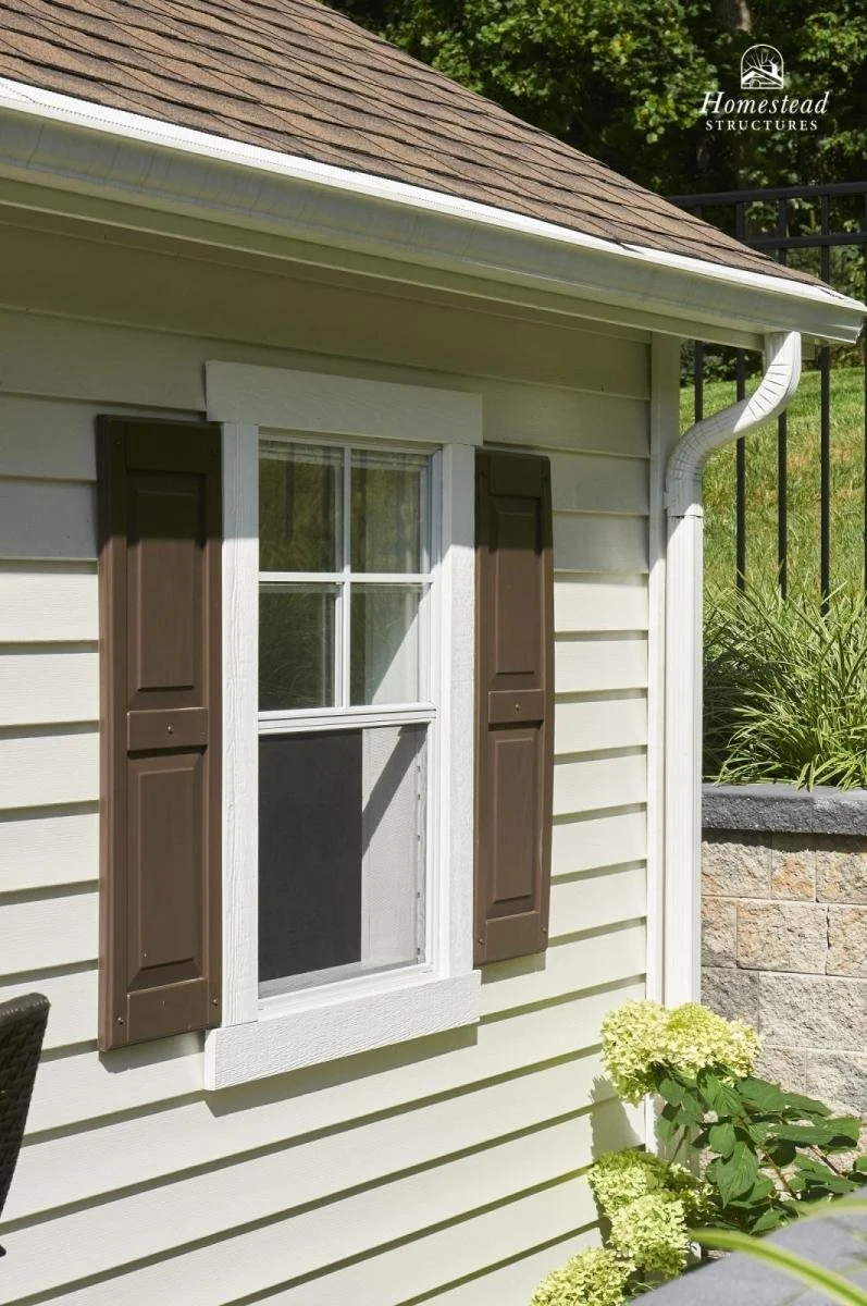 Close-up of a house exterior showing a double-hung window with white trim and dark brown shutters, beige vinyl siding, a roof with brown shingles, a white gutter and downspout, and a garden with green plants and light-colored hydrangea flowers.