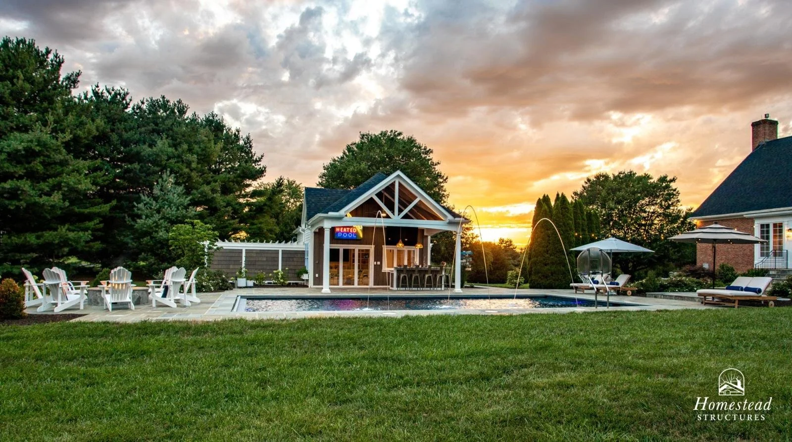 Backyard with a pool, outdoor seating, a pool house with a sign that says Heated Pool, and sunset in the background.
