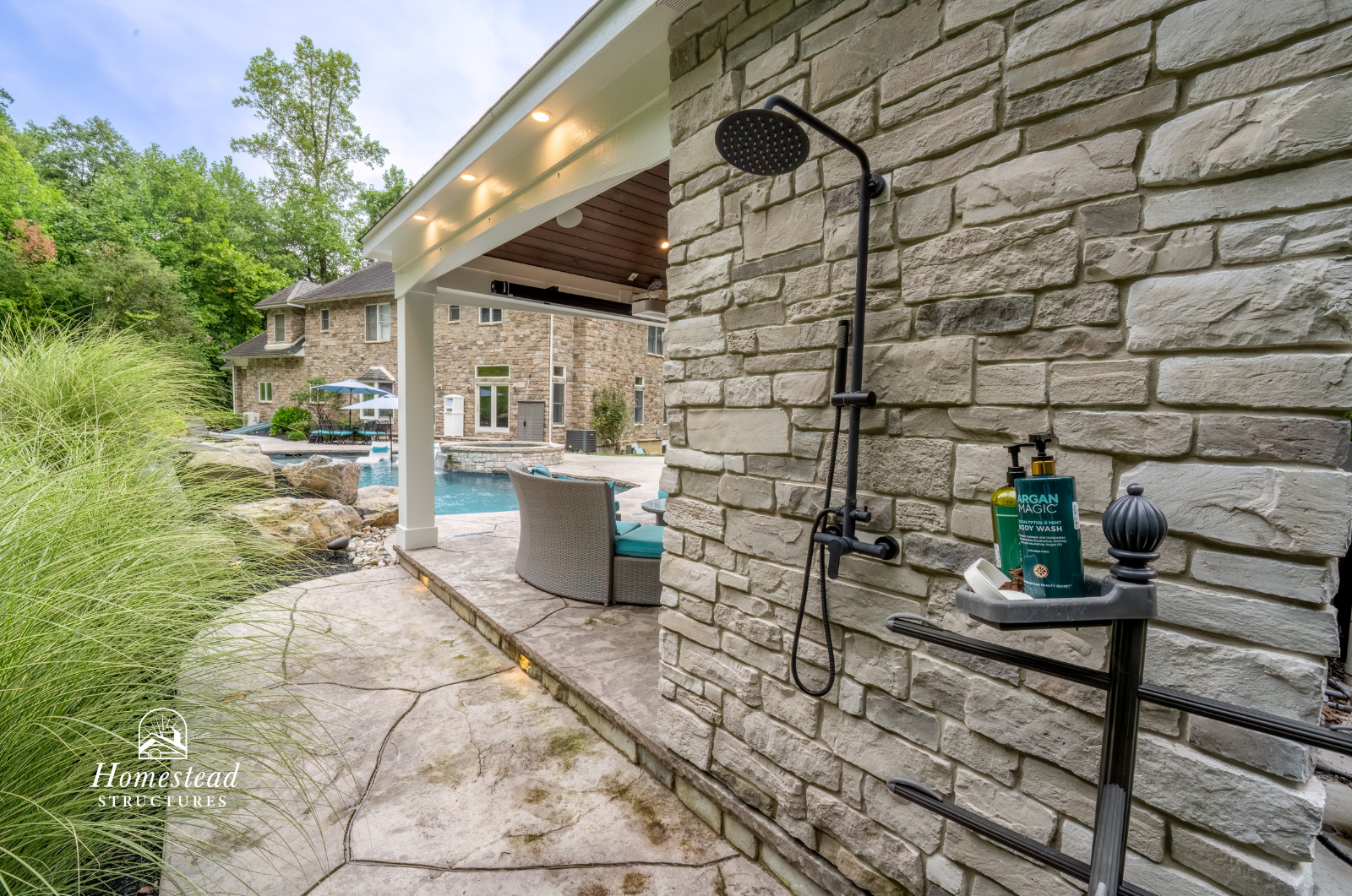 Outdoor patio featuring a built-in shower with stone wall, seating area with cushioned chairs, swimming pool, and a multi-story brick house in the background, surrounded by greenery.