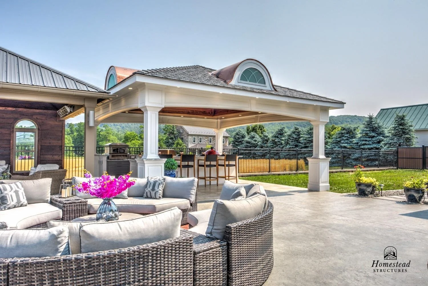 Outdoor patio with seating area, flowers, a built-in bar with bar stools, and a covered pavilion with architectural details. A fence surrounds the area, with trees and a house in the background.