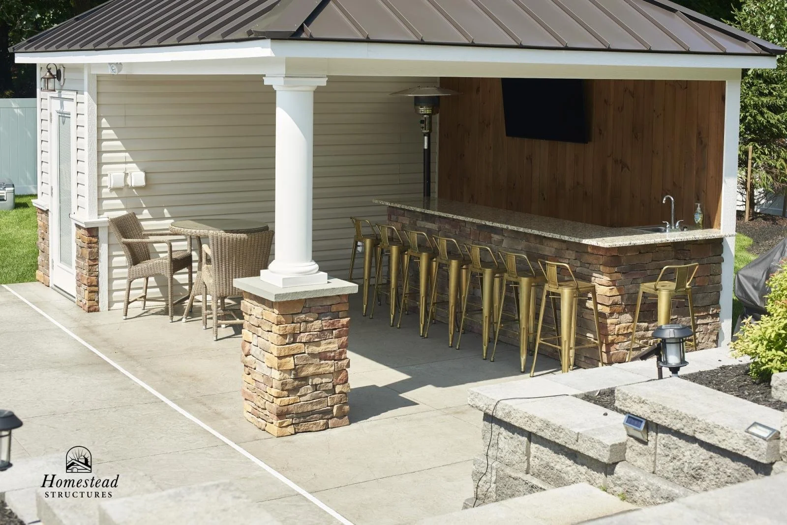Outdoor bar area with a granite countertop, barstools, and a TV mounted on a wooden wall, adjacent to a seating area with wicker chairs and a small round table, on a concrete patio with landscaped surroundings.