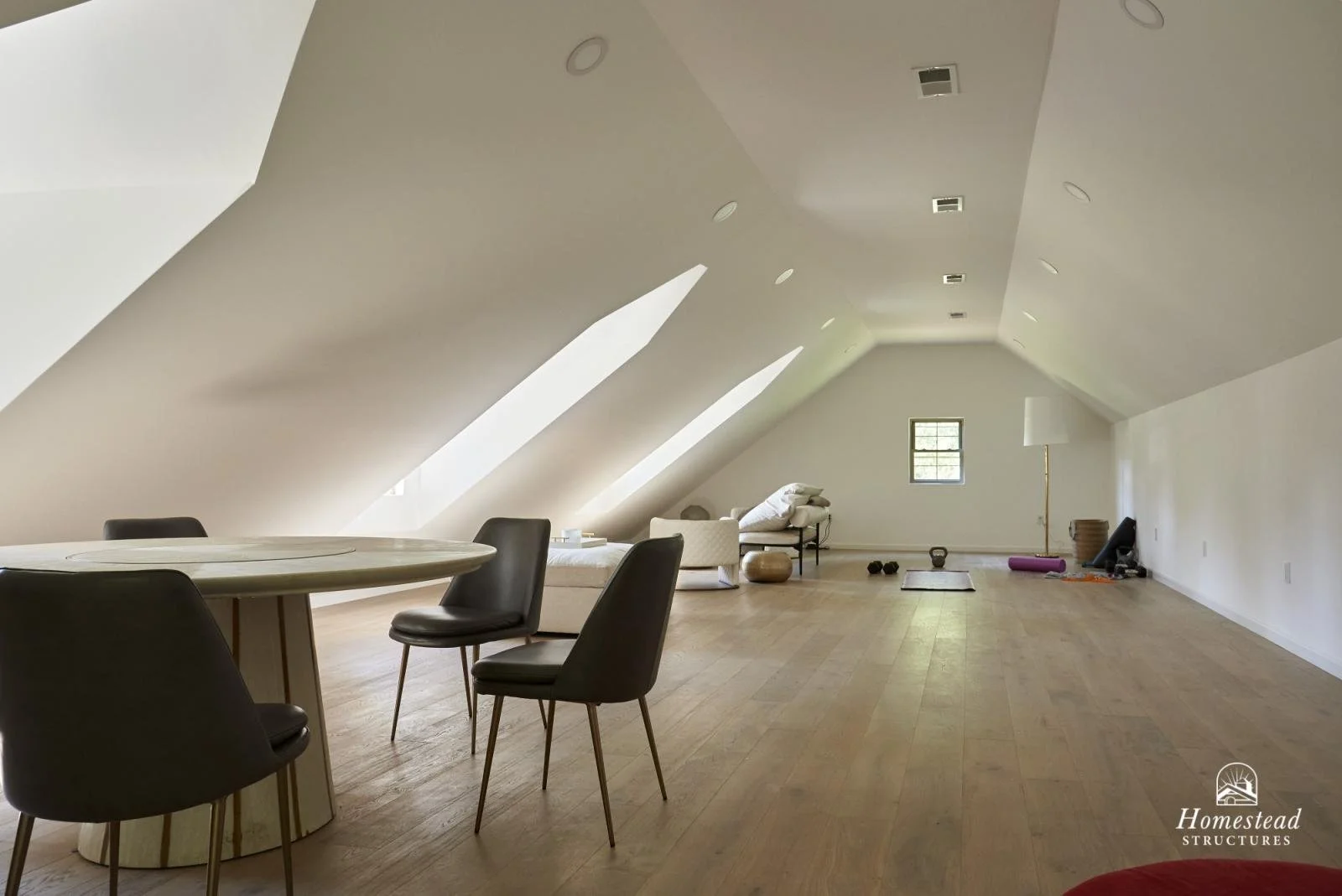 Spacious attic room with sloped ceiling, hardwood floors, and minimal furnishings including a round table with four chairs, a white armchair, a floor lamp, and a small window. The room appears to be used for exercise or relaxation.