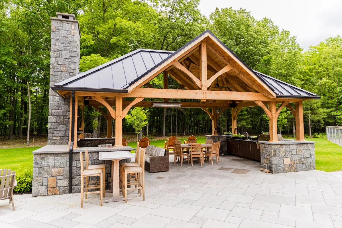 An outdoor covered patio with wooden beams, stone pillars, and a metal roof, featuring built-in outdoor kitchen amenities and seating, surrounded by green trees and lawn.