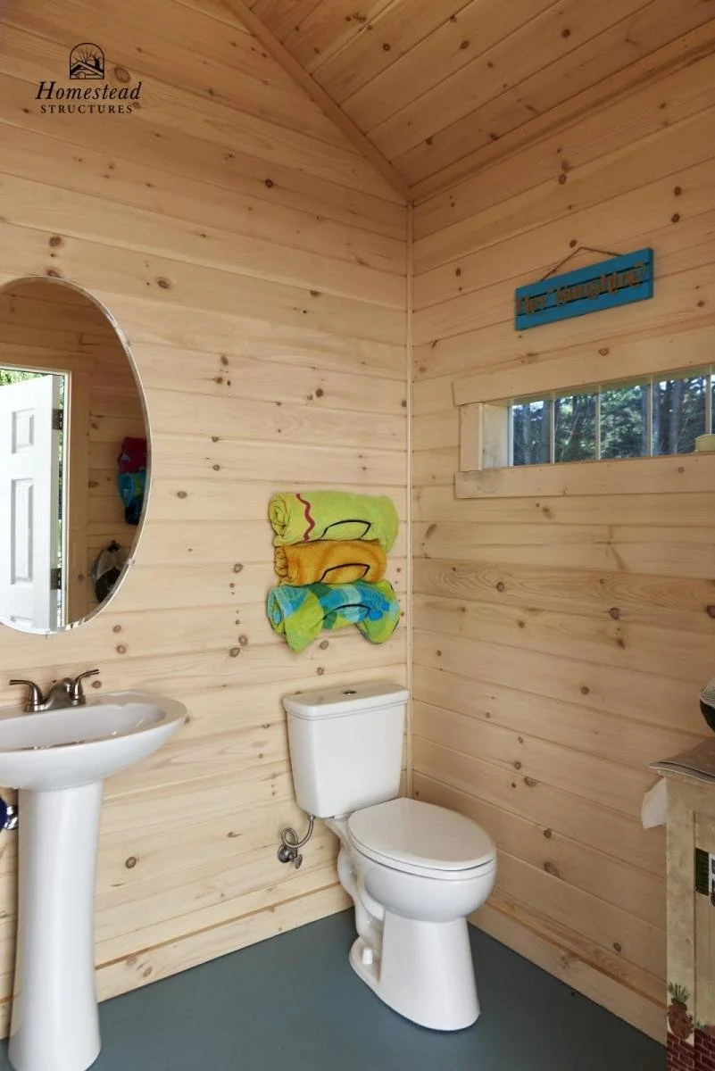 A small bathroom with light-colored wooden walls and ceiling, a white toilet, a white pedestal sink, a round mirror, and colorful towels hanging on the wall. There is a small high window and a decorative sign that reads 'Hickory Ridge'.