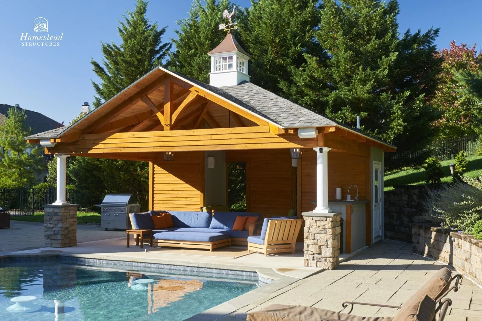 A backyard with a swimming pool, outdoor seating, a wooden pavilion with a roof, trees, and a small cupola on top of the pavilion.