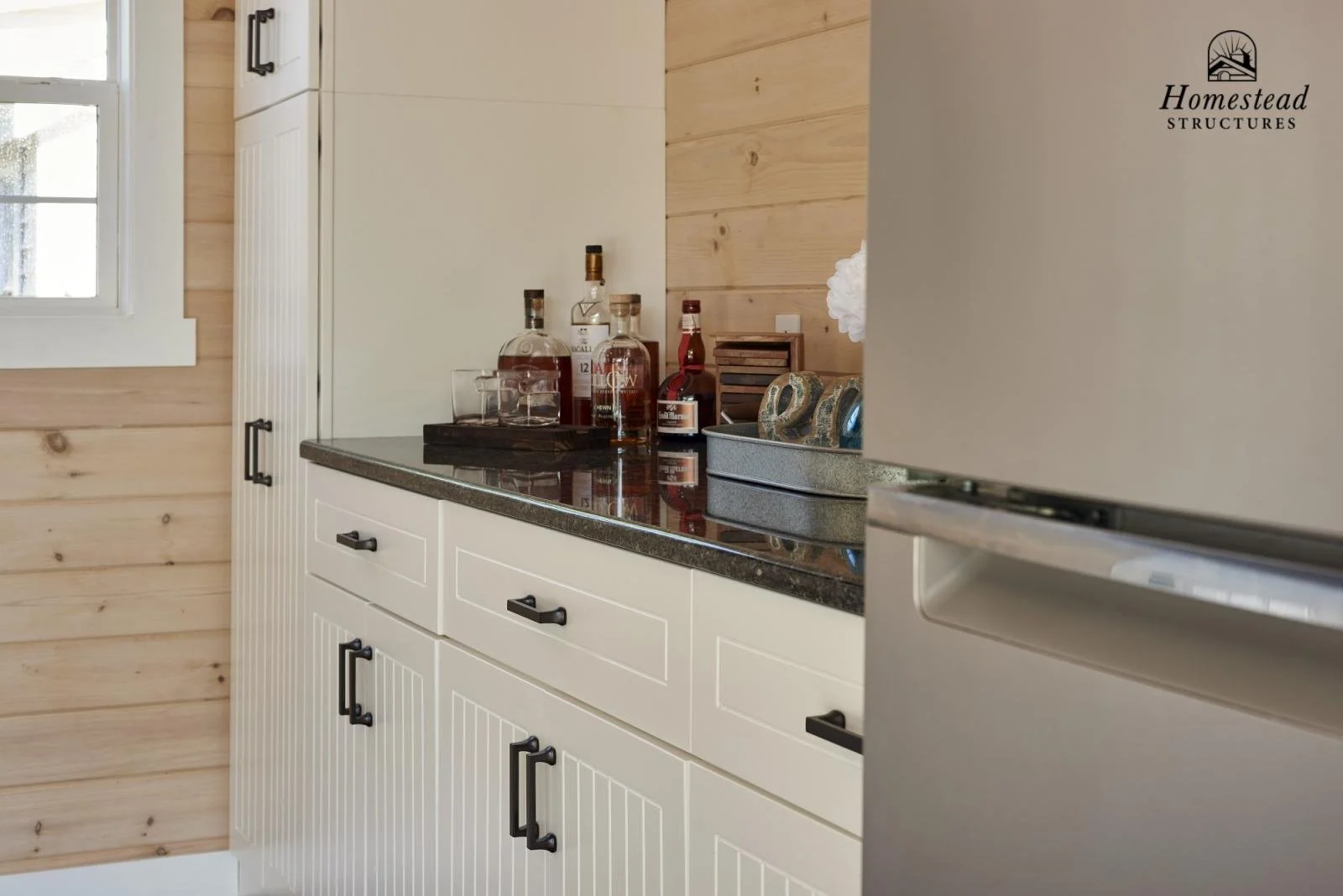 Kitchen with white cabinetry, black handles, a black countertop, bottles of alcohol, glassware, a gray storage box, and a wooden wall panel near a window.