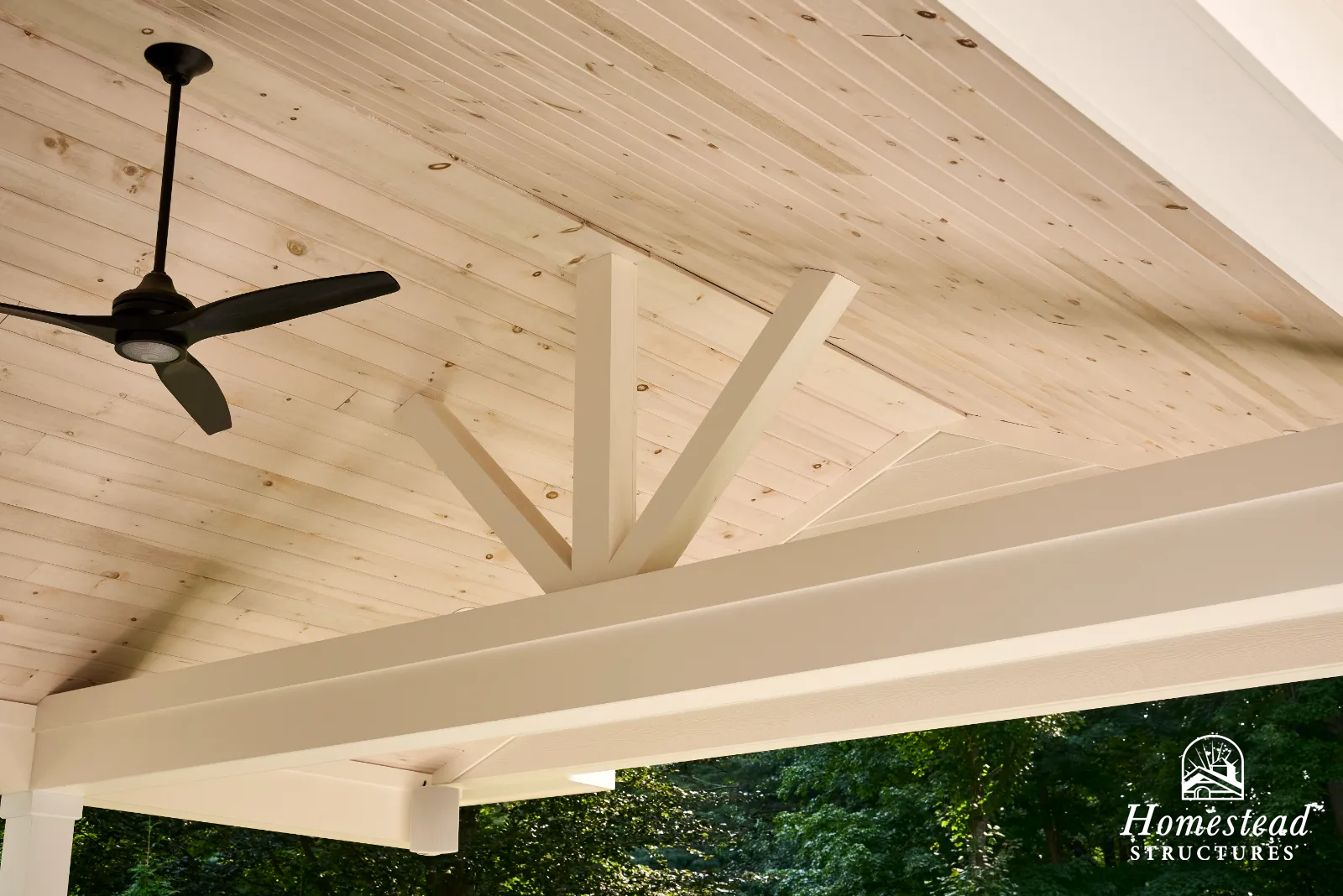 Close-up of a wooden outdoor porch ceiling with a black ceiling fan and white wooden beams, with trees in the background.