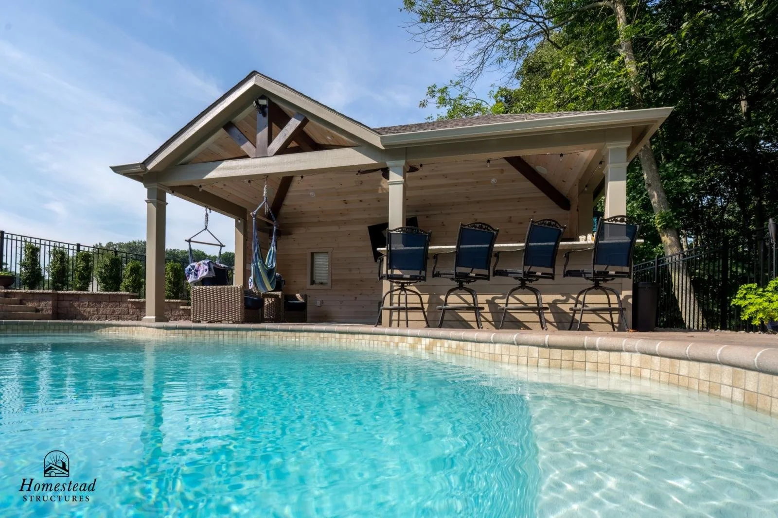 Backyard pool with a shaded poolside lounge area featuring bar stools and hanging hammock chairs, surrounded by greenery and a black metal fence.
