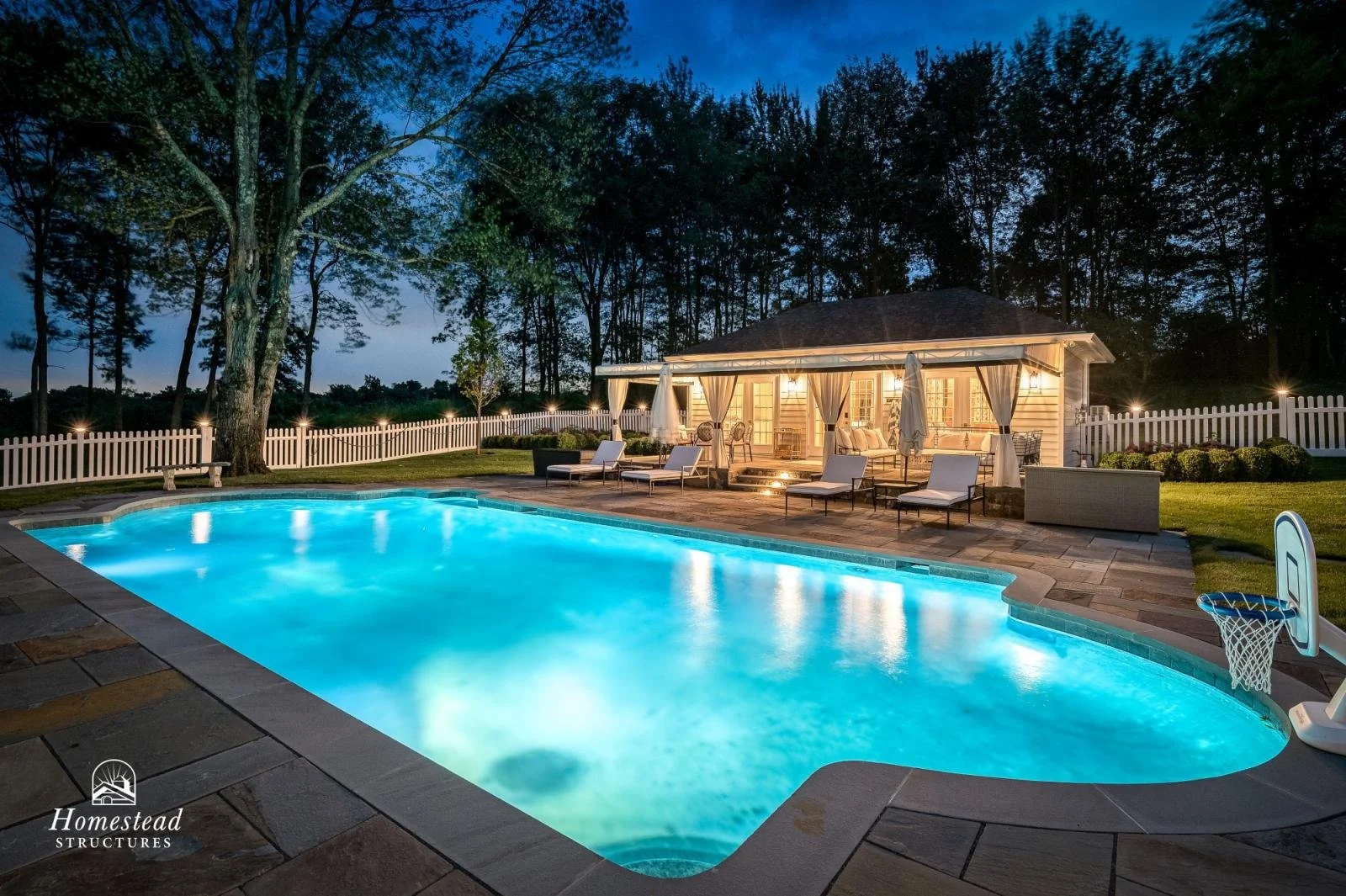 Nighttime view of a lit swimming pool in a backyard, with a house and patio with outdoor furniture, surrounded by trees and a white picket fence.