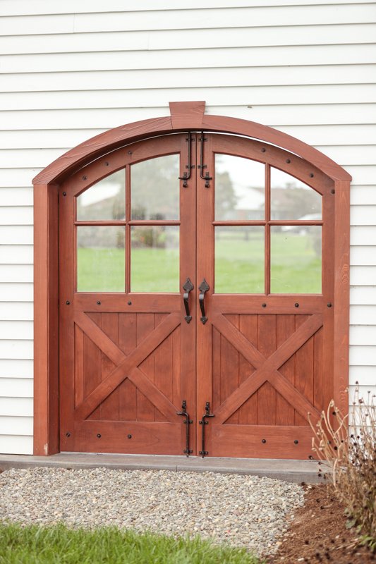 Red wooden barn-style double doors with window panes, installed on a white building exterior, with a gravel pathway in front.