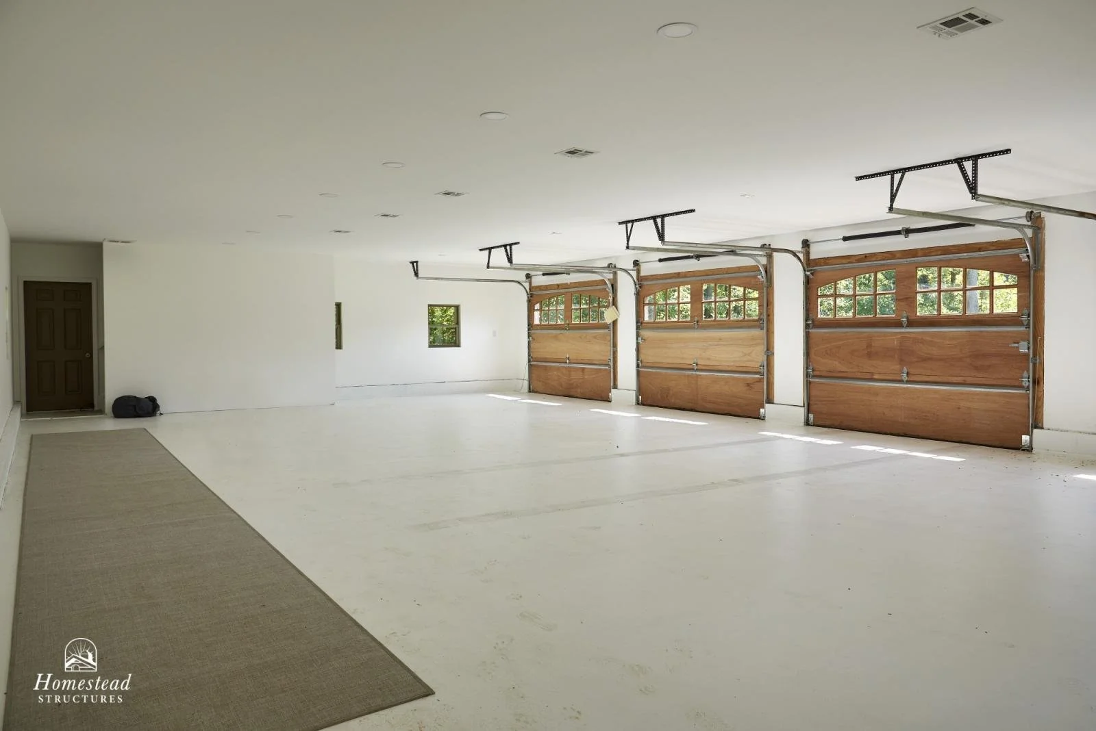 Empty two-car garage with three wooden garage doors, small window, and a brown door in the corner, with a beige rug on the concrete floor.