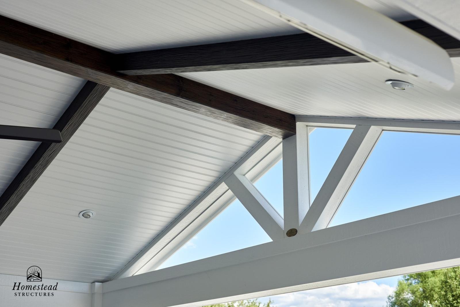 Interior view of a house ceiling with white and dark wood beams and a large triangular window showing a blue sky and trees outside.