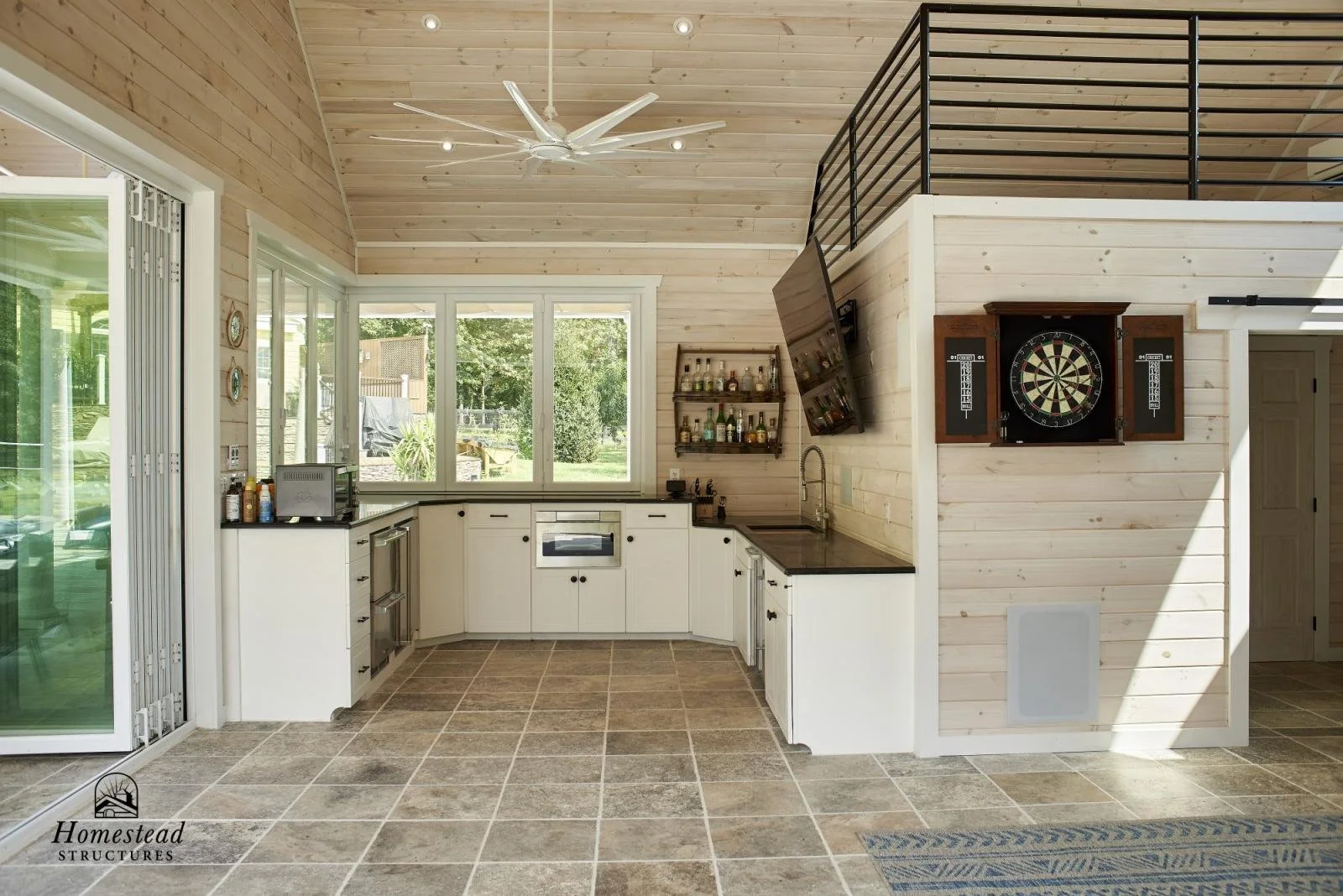 Interior of a home kitchen with large windows, white cabinets, black countertops, and a dartboard on the wall, with a partial view of outdoor greenery through the windows.