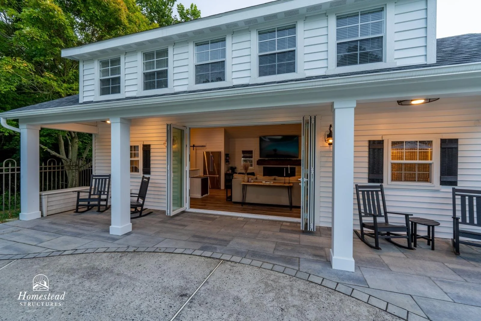 View of a white two-story house with a covered patio area outside, shaded by trees, with black rocking chairs and an open sliding door revealing a living room inside.