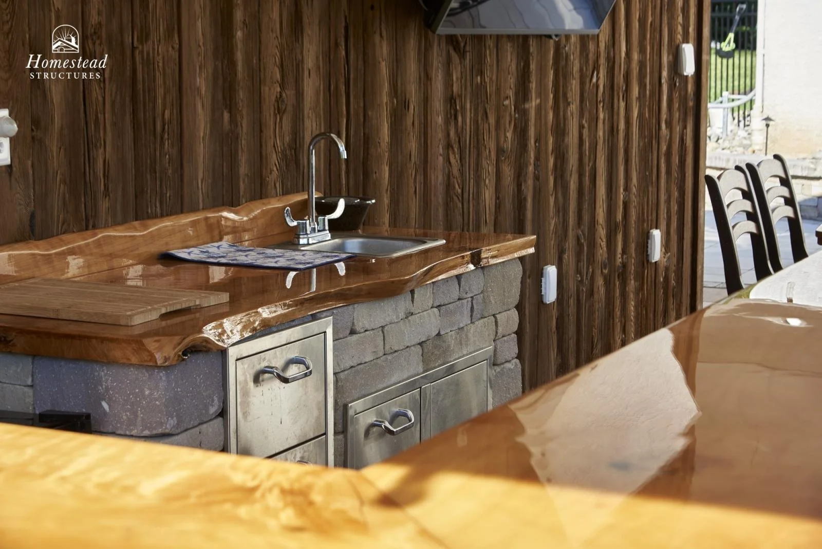 Outdoor kitchen area with a wooden counter, stainless steel sink, and a wooden wall background. There is a chopping board, dish towel, and part of a dining table with chairs visible.