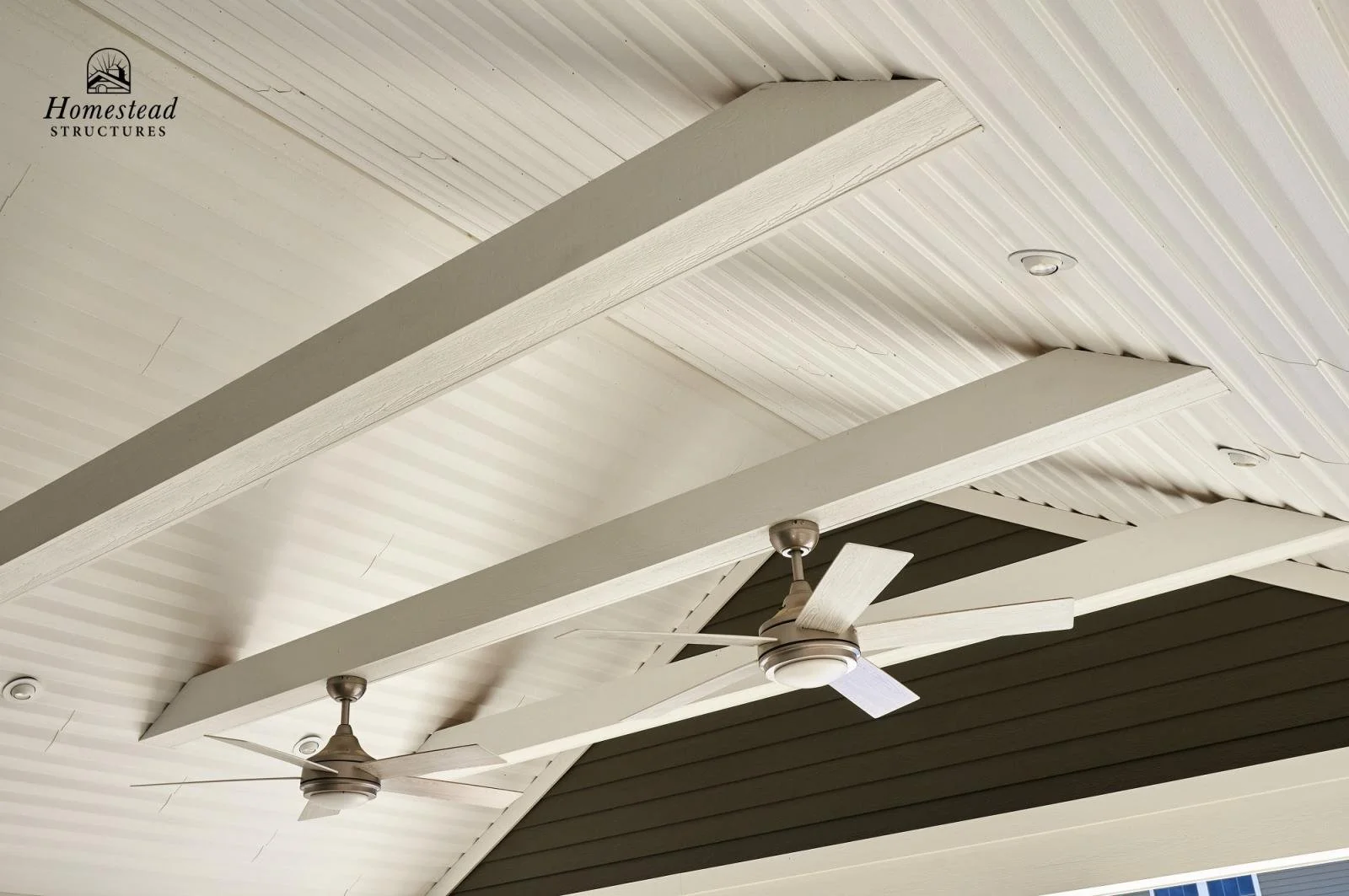 The ceiling of a porch or patio area with two modern ceiling fans and decorative beams, with a side wall below painted in dark brown.