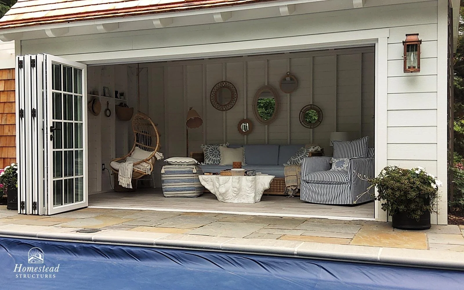 Interior of a pool house with a seating area, wicker hanging chair, striped armchair, sofa, and wall mirrors, with an open foldable door.