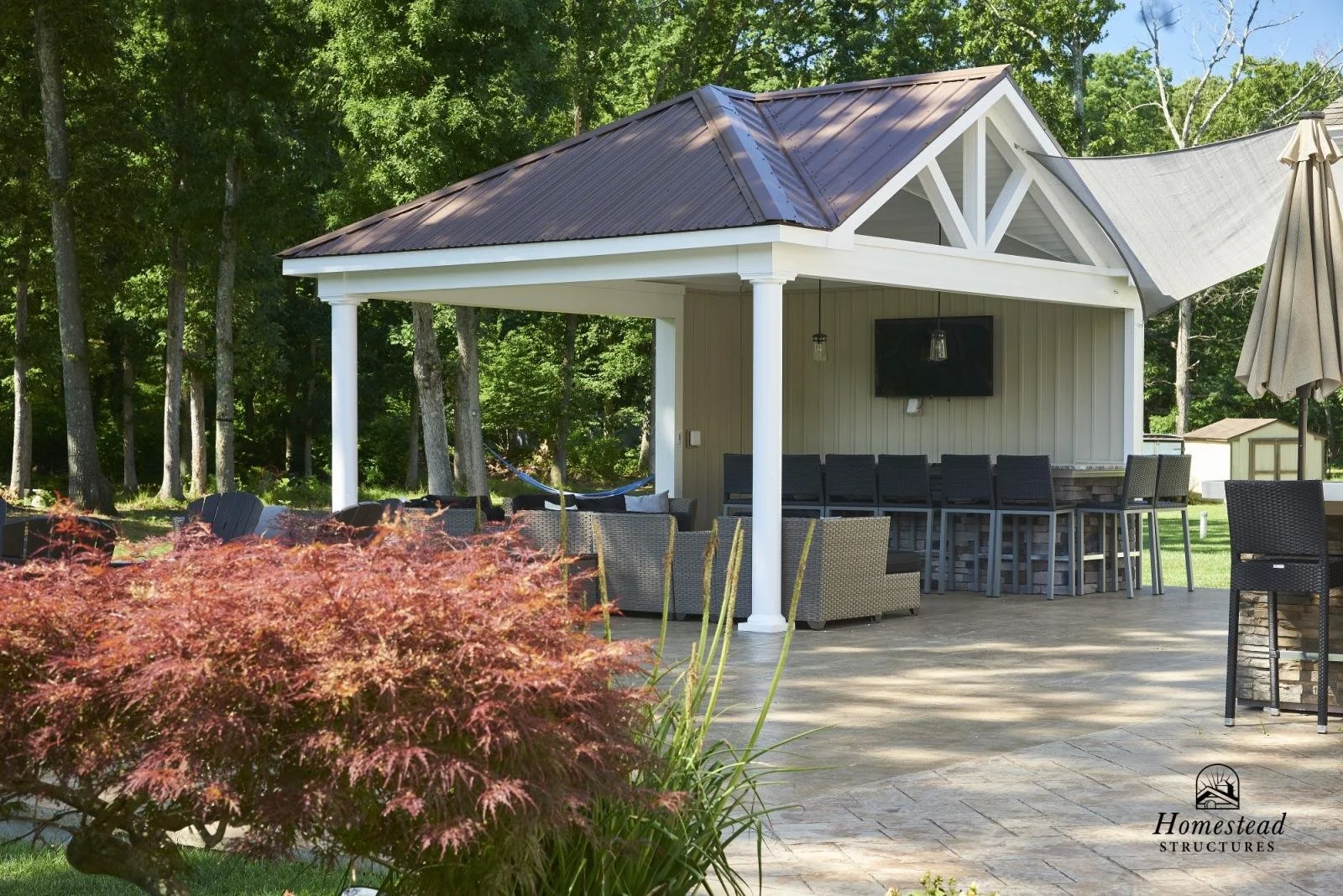 An outdoor patio area with a covered bar and seating, surrounded by trees and landscaped with bushes, during the daytime.
